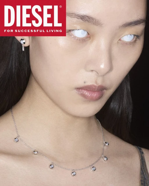 Portrait of a young woman with glowing white eyes wearing a silver necklace and Diesel earring in a dimly lit photo studio.