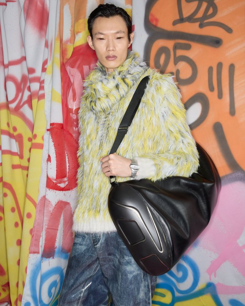 An Asian man in a fur-style sweater and jeans poses with a large black bag in front of a graffiti-covered wall. Fashion shot.