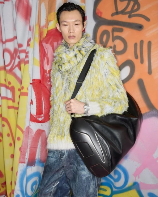 An Asian man in a fur-style sweater and jeans poses with a large black bag in front of a graffiti-covered wall. Fashion shot.