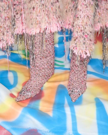 Fabric sculpture of high heel shoes with a fuzzy skirt and colorful graffiti background in shades of pink, brown, blue, yellow, and white.