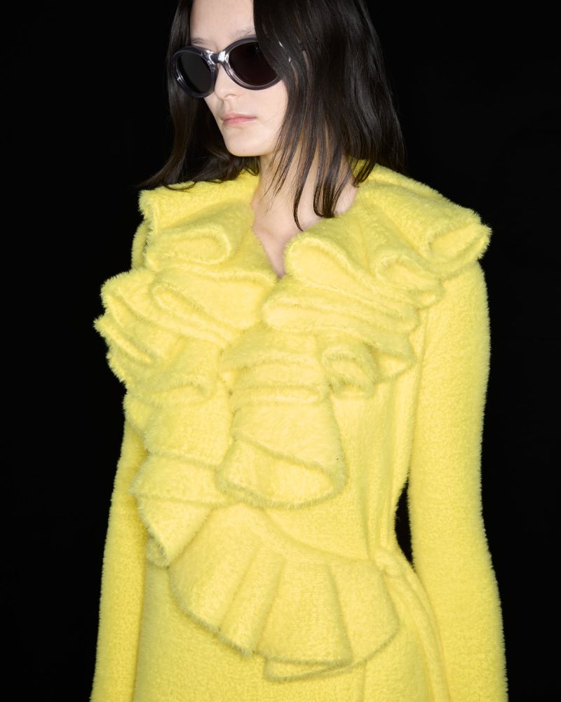 Woman with dark hair wearing sunglasses and a bright yellow coat with ruffles against a dark background in a close-up studio shot.