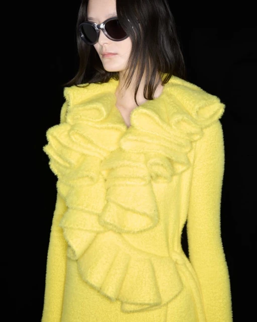 Woman with dark hair wearing sunglasses and a bright yellow coat with ruffles against a dark background in a close-up studio shot.