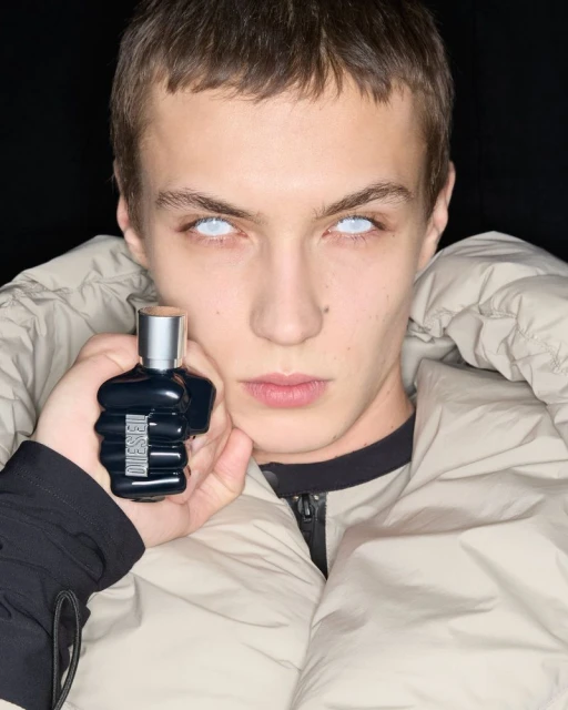 Close-up of a young adult man holding a Diesel fragrance bottle, wearing a tan puffer jacket against a dark background.