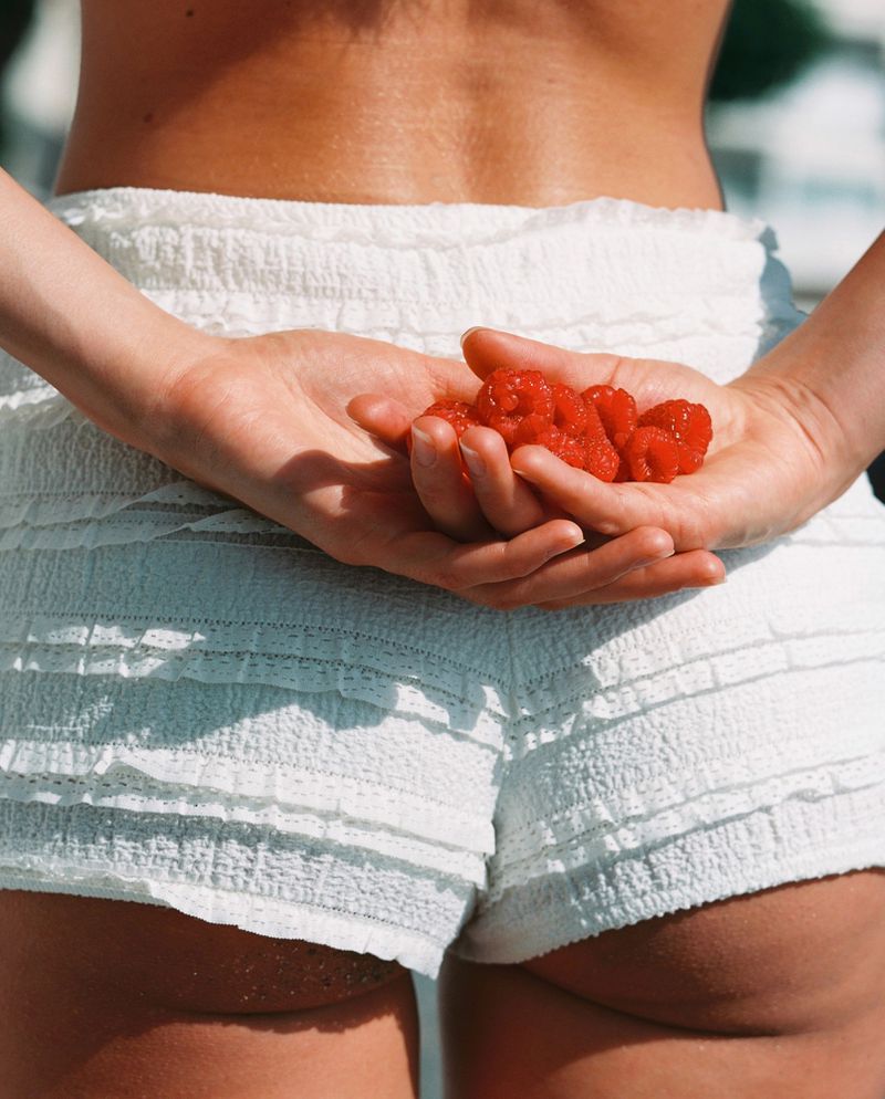 Back view of a person in white shorts holding a handful of ripe red raspberries, with light blue background, bright studio lighting.
