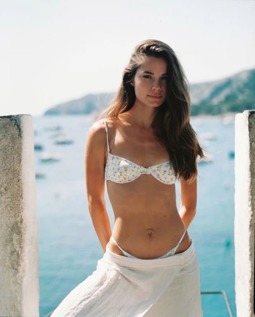 Attractive woman in bikini and white wrap standing outdoors near ocean with sailboats on a sunny day, looking at the camera.
