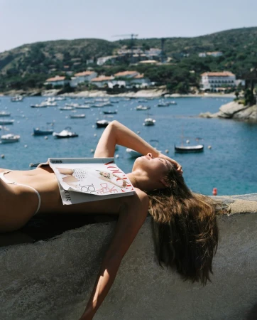 Woman lying on stone wall by the sea with a magazine over her chest, shot by Maurizio Pedroni.