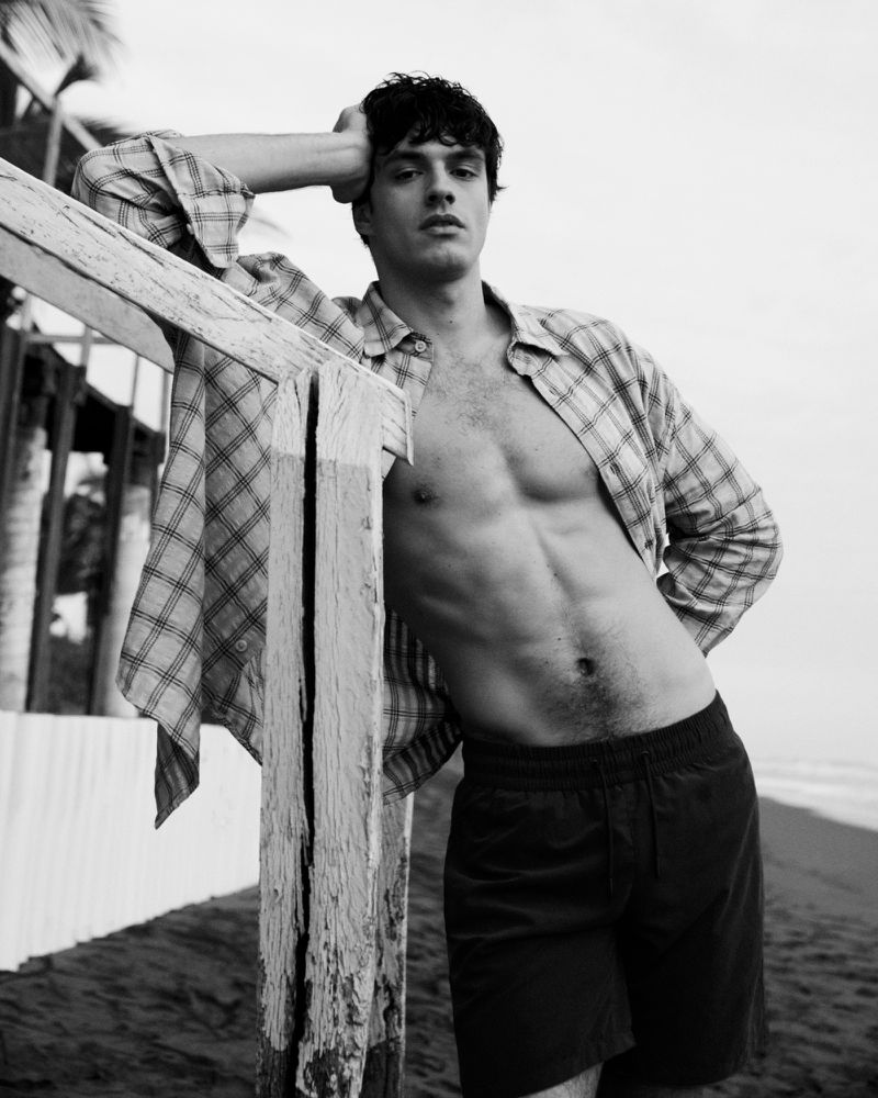 A tanned male model poses shirtless in a plaid shirt and dark shorts, leaning against a rustic wooden structure on a sandy beach.