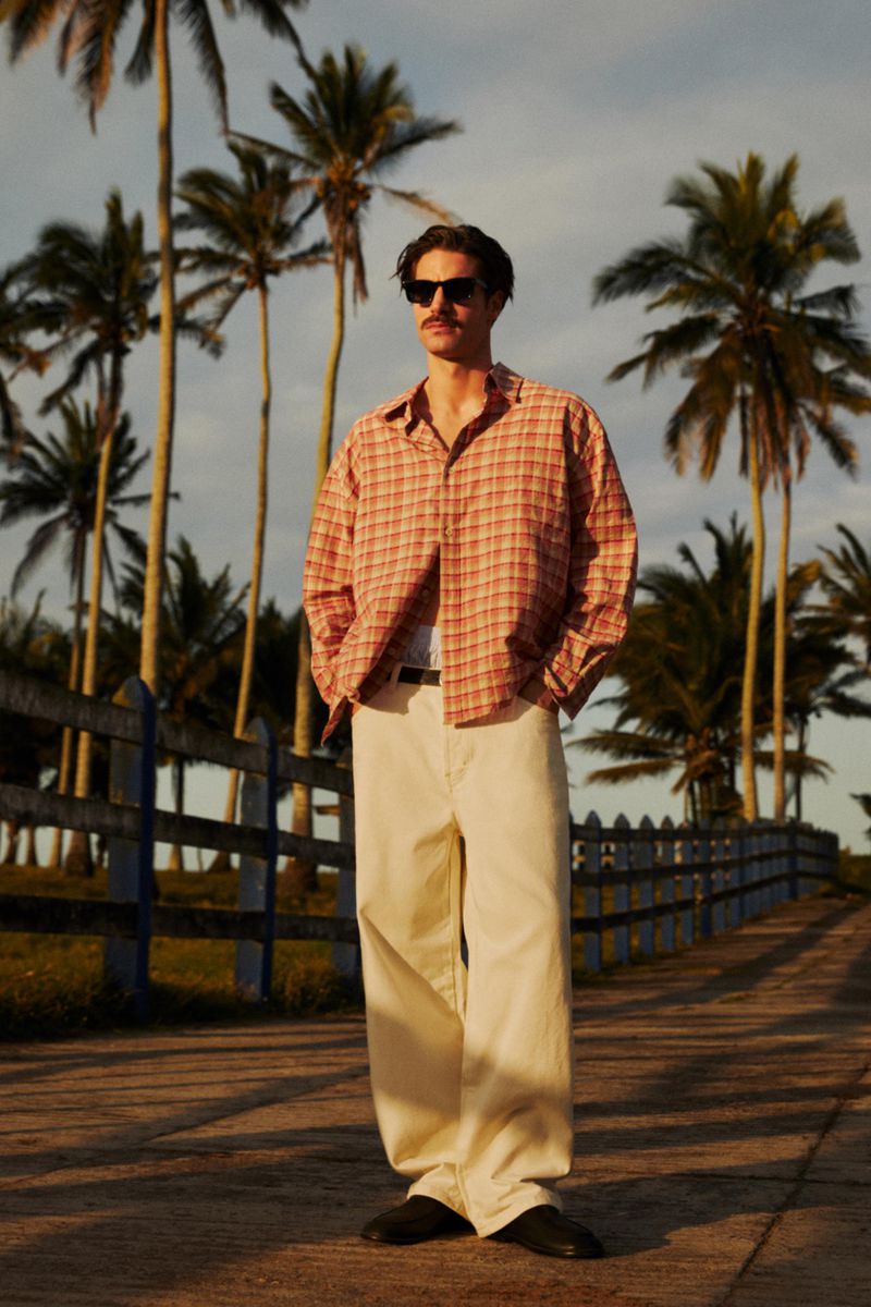 A fair-skinned adult man with sunglasses poses in a patterned shirt and tan pants with palm trees and a blue fence background.