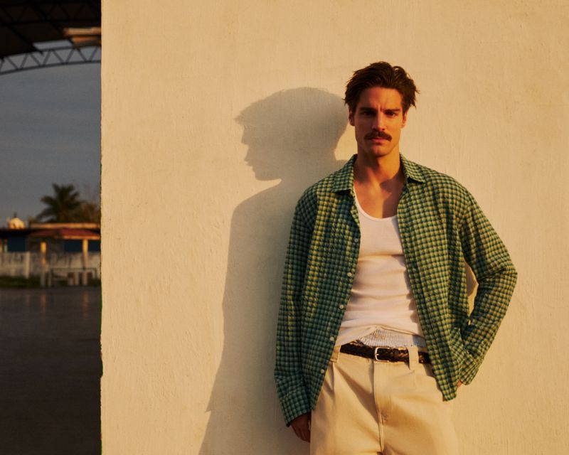 Man with a mustache poses outdoors wearing an unbuttoned green plaid shirt and white tank top against a white wall in golden light.