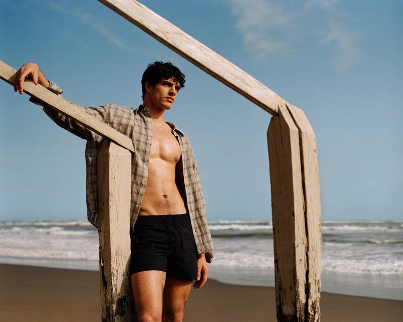 Male model in an open plaid shirt and dark swim trunks leaning against a wooden structure on a sandy beach.