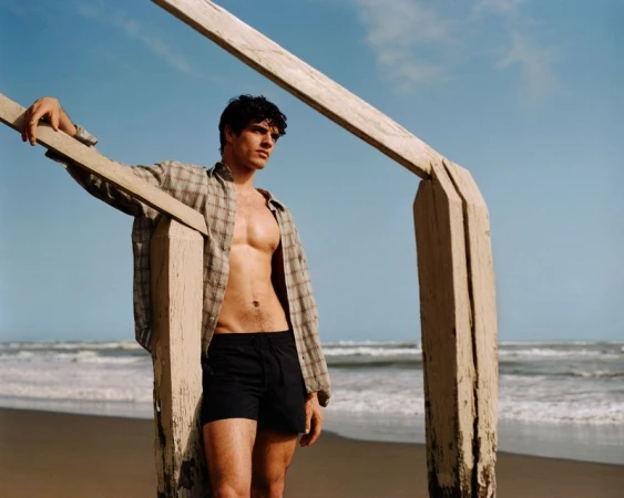 Male model in an open plaid shirt and dark swim trunks leaning against a wooden structure on a sandy beach.