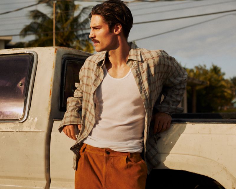 Man in a white tank top and plaid shirt leans against an old truck, sporting brown trousers, with an outdoor, daytime setting.