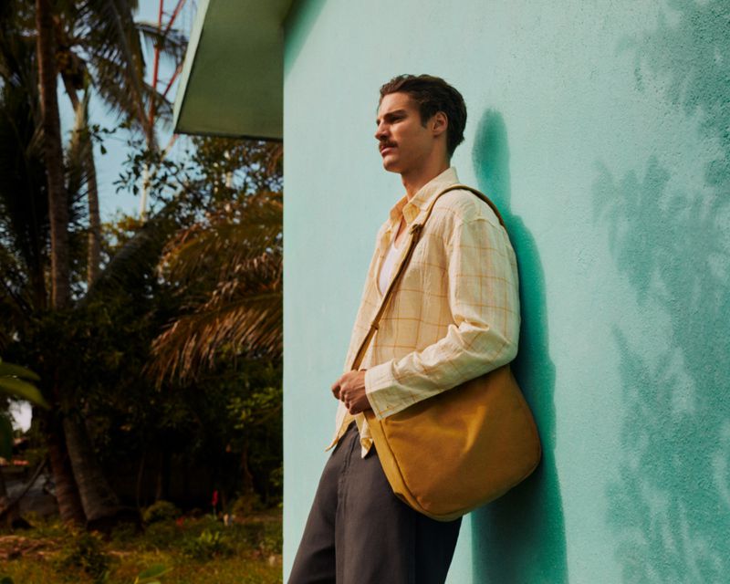 Adult male model leans against a light blue wall outdoors, wearing a yellow shirt, dark pants, and yellow satchel, under palm trees.
