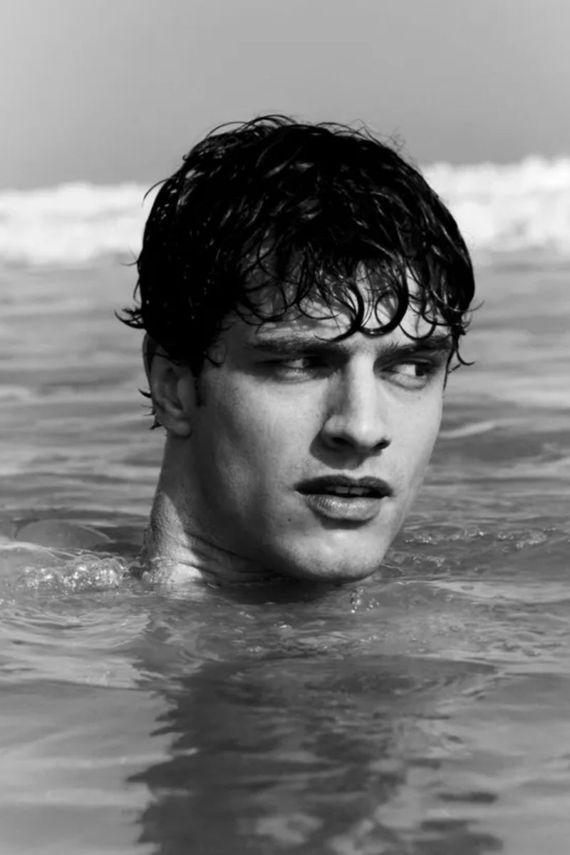 Close-up black and white portrait of a man with wet dark hair submerged in water, looking off to the side.