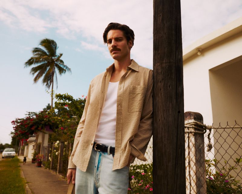 Man with a mustache leans against a post in a casual outfit featuring a patterned shirt and light wash jeans on a sunny day.