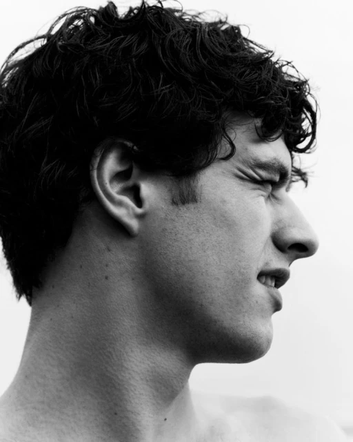 Monochrome portrait of a young adult male with curly dark hair, eyes closed, and a slight grimace, captured in a high-contrast studio setting.