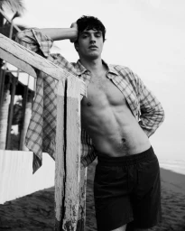 Black and white portrait of a male model leaning against a wooden structure on a sandy beach wearing a plaid shirt and dark shorts.