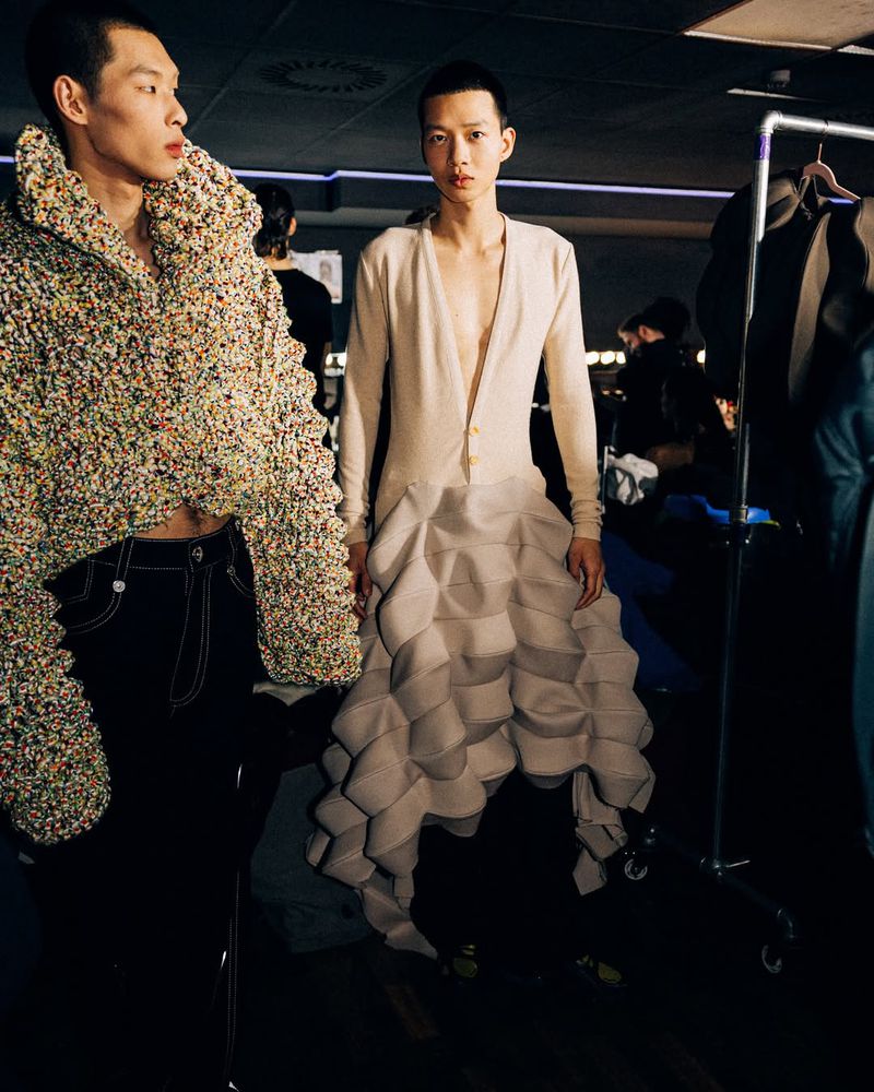 Two runway models pose in avant-garde fashions; one in a textured cropped jacket, the other in a pale cardigan with a geometric skirt.