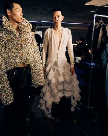 Two runway models pose in avant-garde fashions; one in a textured cropped jacket, the other in a pale cardigan with a geometric skirt.