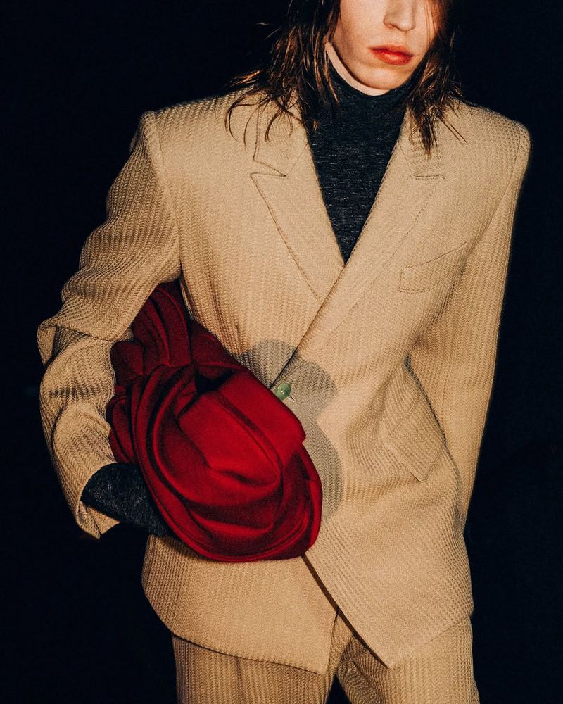 Person in beige suit with a black turtleneck, holding a red textile object. Fashion editorial photography.