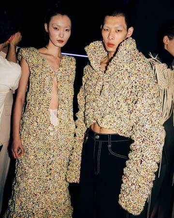 Two fashion models showcase textured, patterned outfits in a dimly lit setting. The garments feature unique textures and colors.