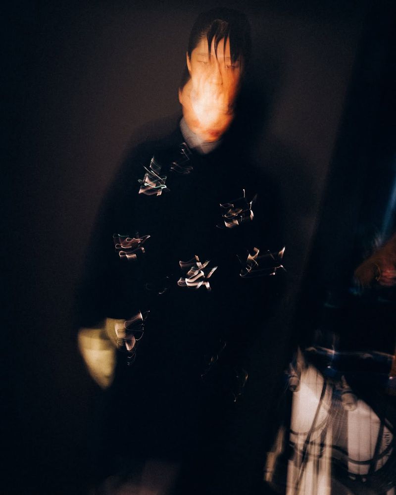 Abstract portrait of a person with blurred face wearing a dark floral shirt, set against a dark background in a blurry, artistic style.