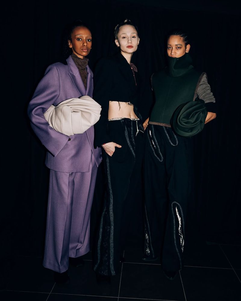 Three fashion models pose indoors against a dark background, showcasing avant-garde clothing with unique textures and designs.