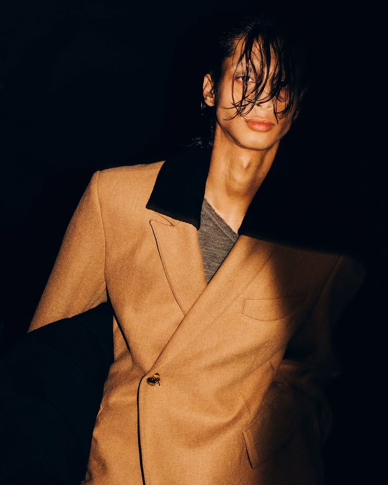 Portrait of an adult male with long dark hair wearing a tan coloured blazer with a black collar against a dark background.
