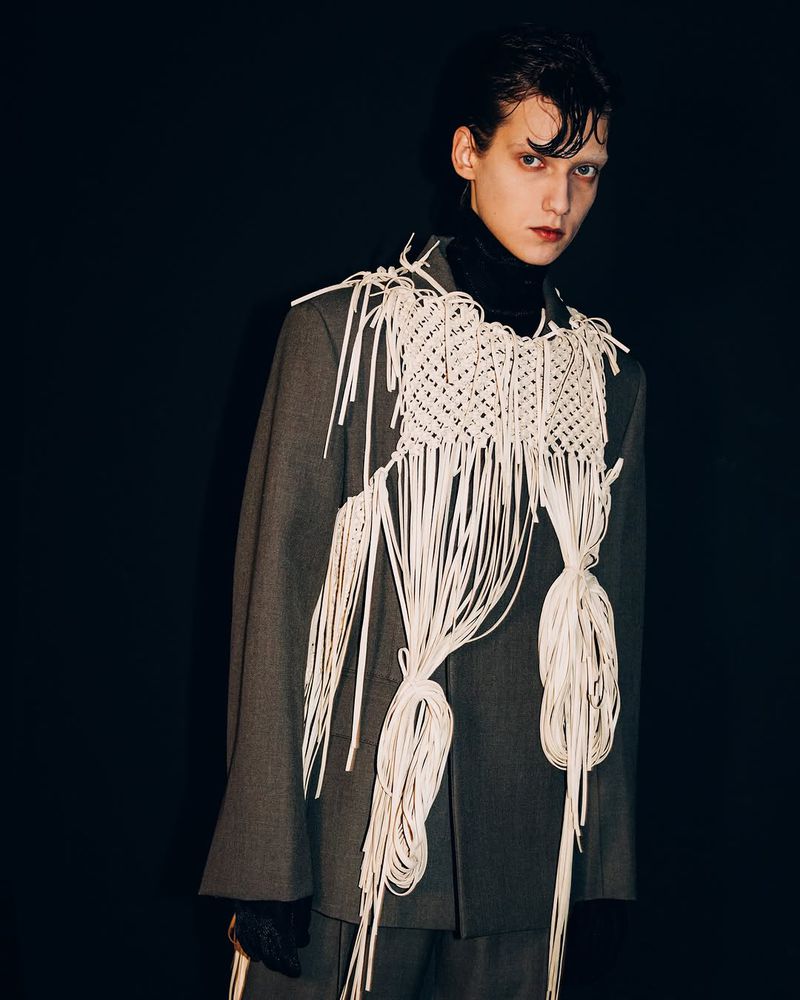 An adult male model with slicked-back dark hair wears a gray suit and a cream-colored macrame-style garment in a studio setting with a dark background.