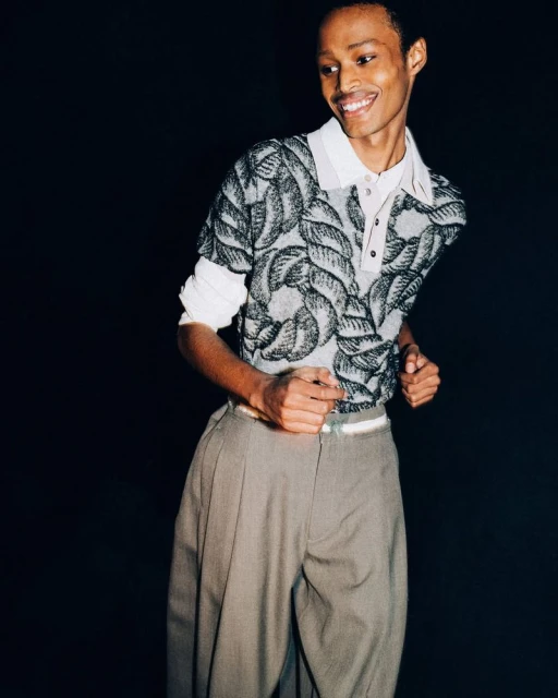 Young man smiles in a fashion portrait wearing a collared sweater with gray pants against a dark backdrop in an indoor setting.