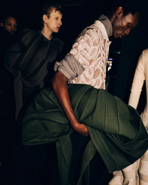 Fashion models in structured green and white outfits backstage at a runway show, captured in a low-light, indoor environment with a shallow depth of field.