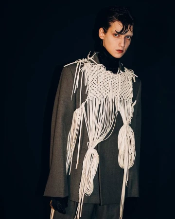 Model wearing a grey blazer layered with an intricate white macrame fringe harness backstage at Pronounce F/W25 in Milan.