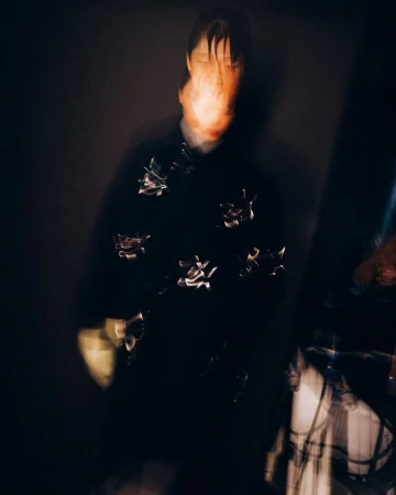 Motion-blurred portrait of a person in a dark coat with metallic accents against a dark backdrop