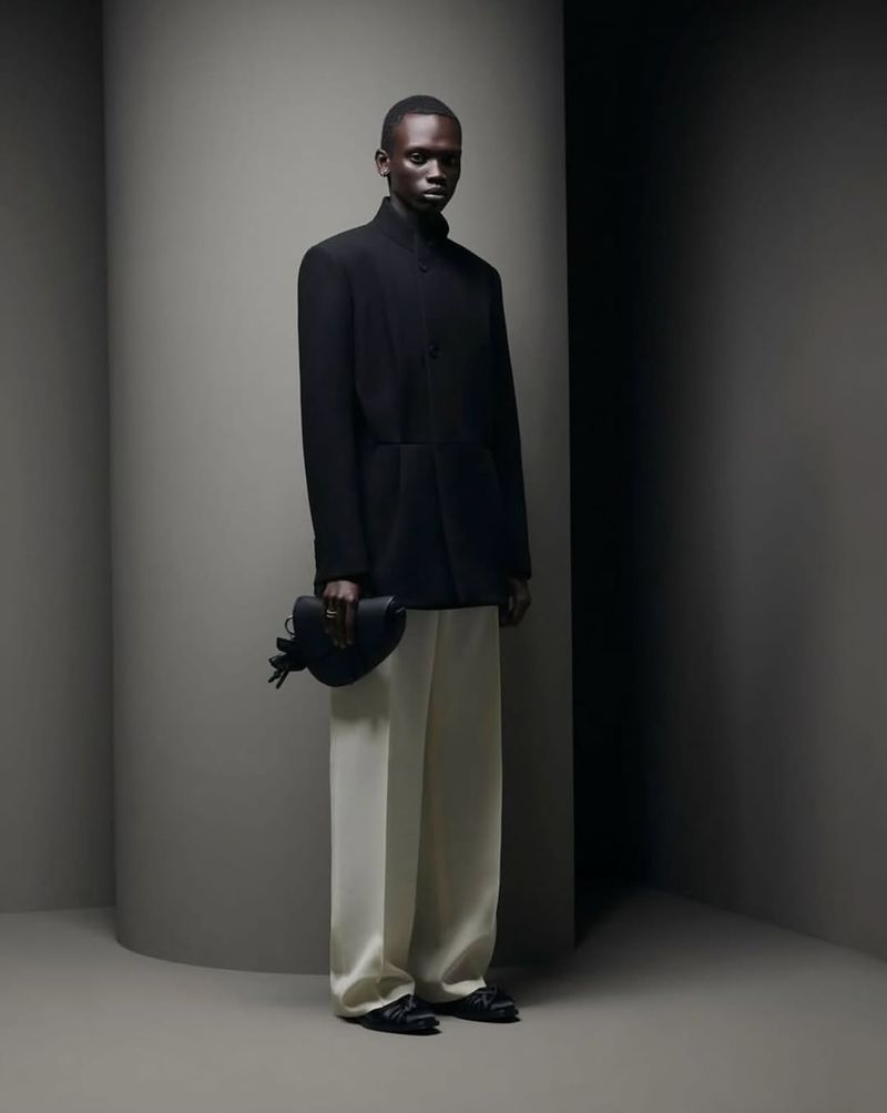 An African male model poses in a black jacket and white pants, holding a small black bag against a grey wall, showcasing a modern and elegant style.