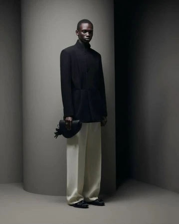 An African male model poses in a black jacket and white pants, holding a small black bag against a grey wall, showcasing a modern and elegant style.