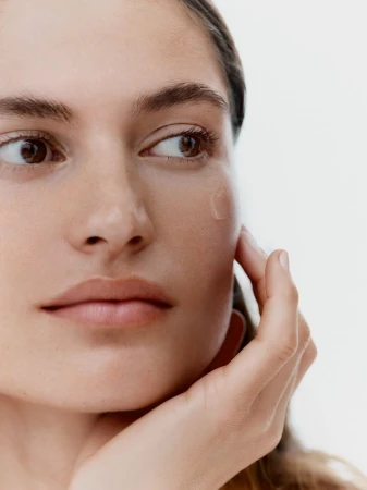 Close-up portrait of a woman with light skin, hand near face, and clear serum visible on her cheek in a bright, neutral setting.