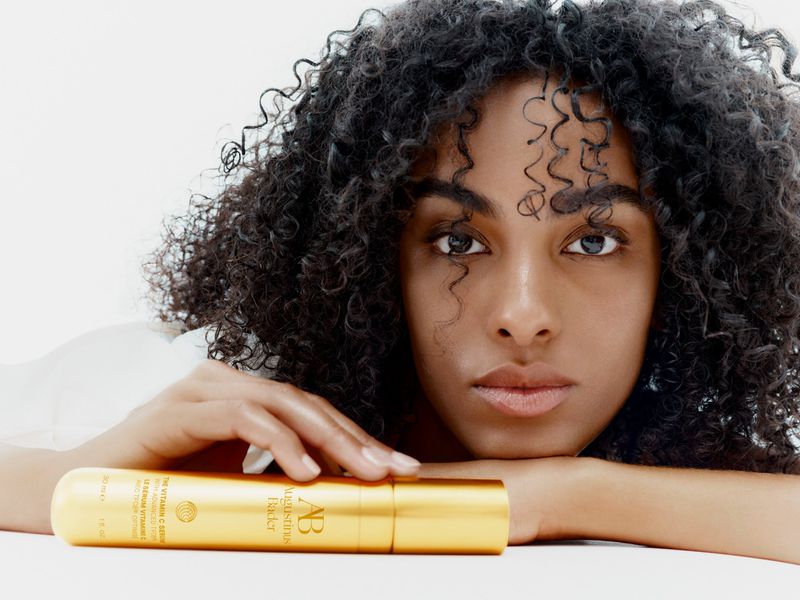 Close-up beauty shot of an adult female with curly hair resting her head on her arms next to a gold skincare container.
