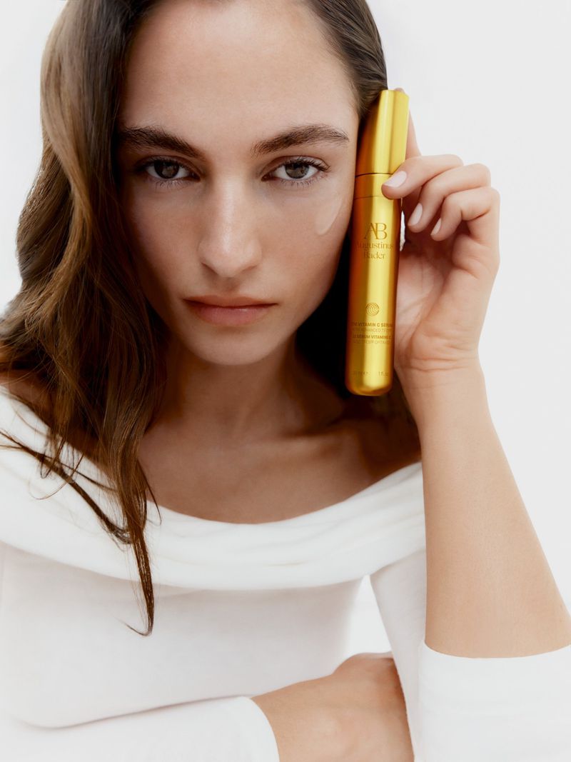 Brunette woman poses with a gold serum bottle in a beauty shot against a white backdrop, showing skincare application.