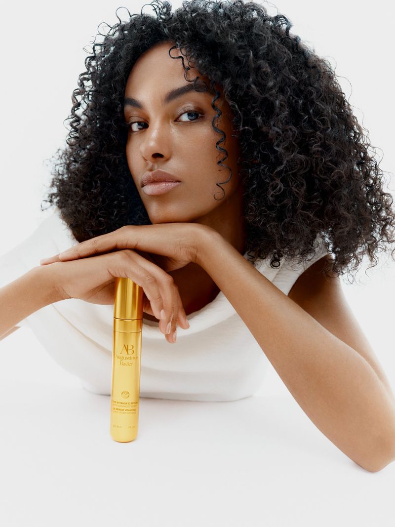 Beauty shot of a Black female model holding a gold skincare product in a studio setting, showcasing natural hair and elegant presentation.