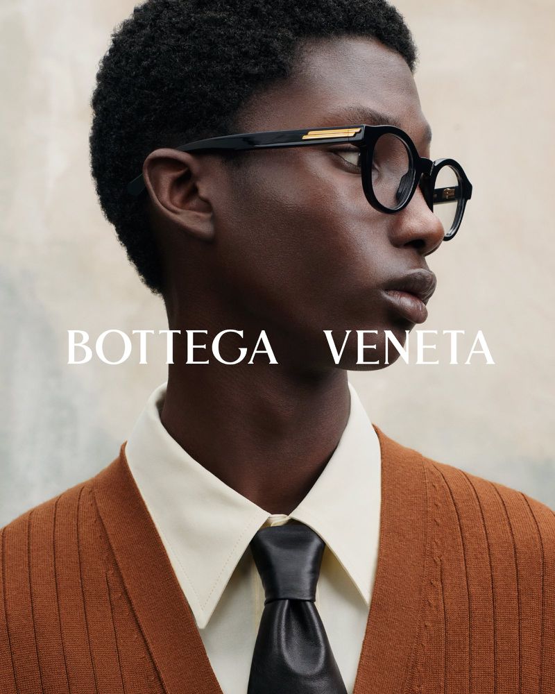 Portrait of a dark-skinned adult man in eyeglasses and a buttoned cardigan, posing against a neutral background, promoting Bottega Veneta.