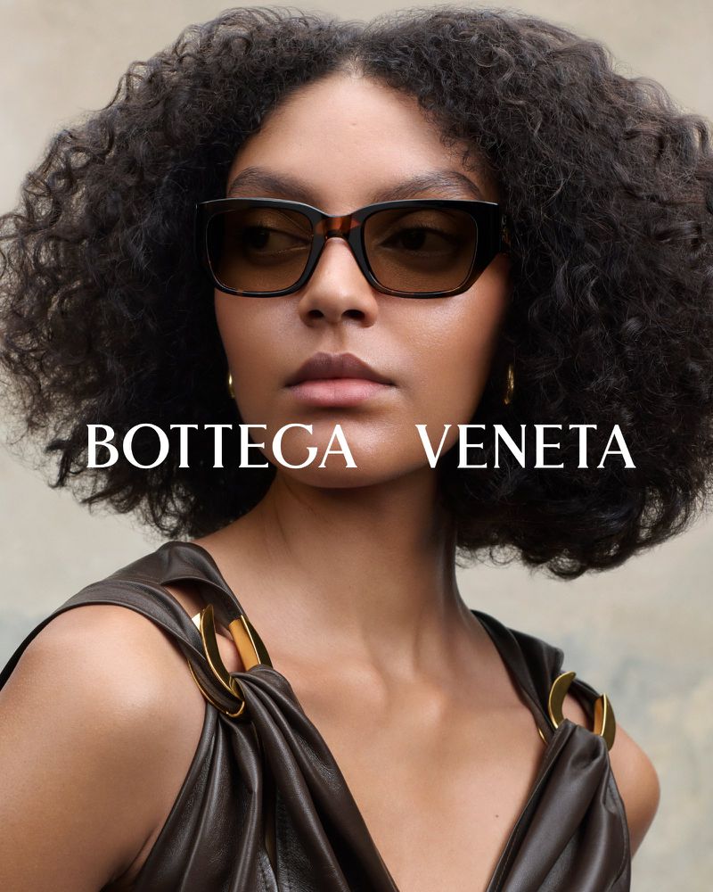 Close-up studio shot of a woman with curly hair wearing sunglasses and a brown dress, posing against a neutral backdrop. Fashion.