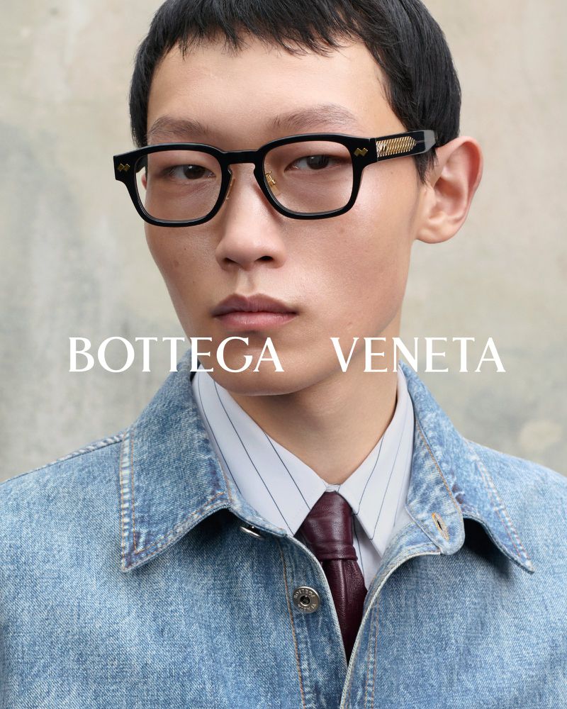 Eyewear fashion shot: a stylish adult male wearing a denim jacket and black-framed glasses, looking directly at the camera.