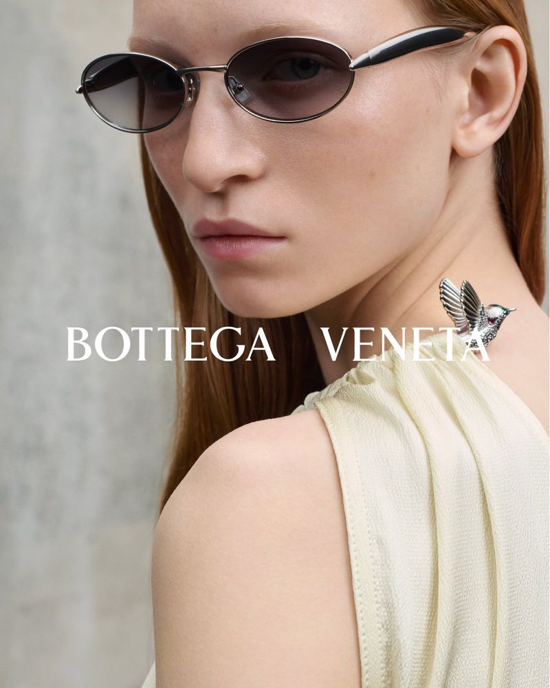 Close-up studio shot of an adult woman wearing sunglasses and a light yellow dress with a bird brooch on her shoulder.