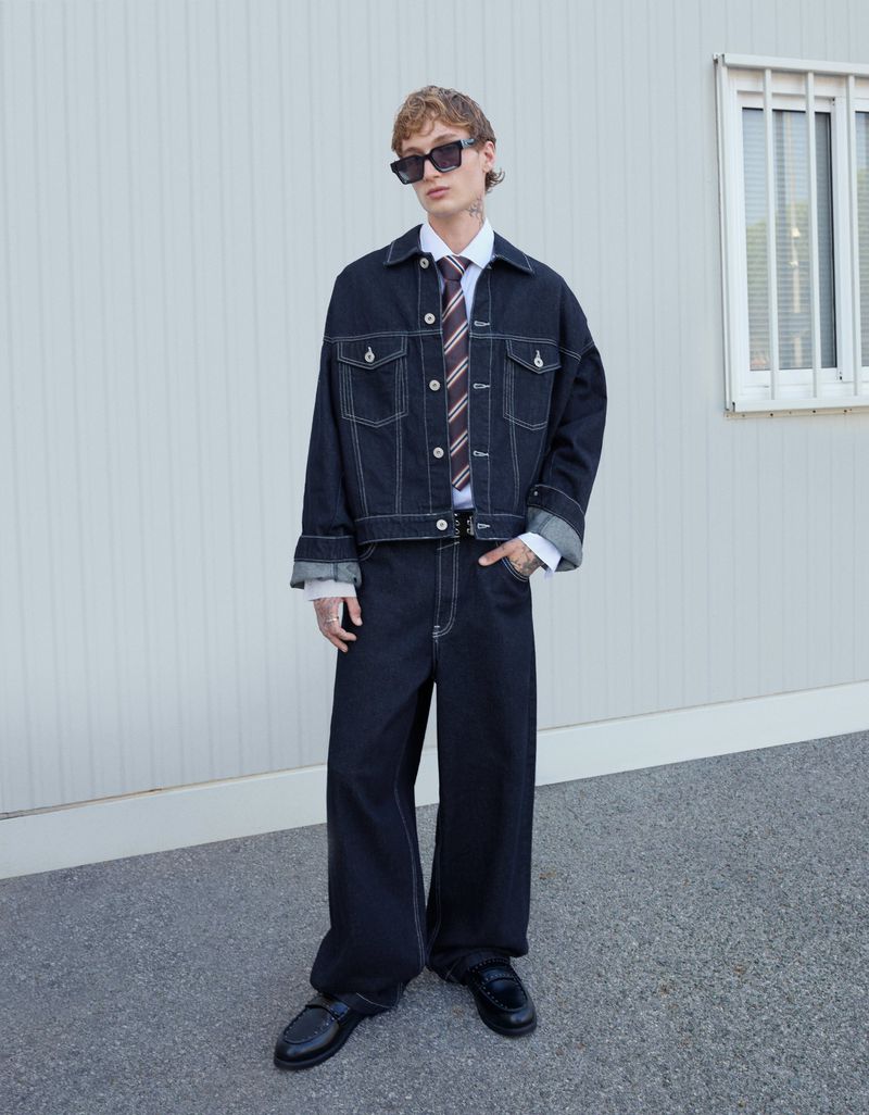 Male fashion model poses in denim jacket and wide-leg jeans with black loafers against a plain wall on an overcast day outdoors.