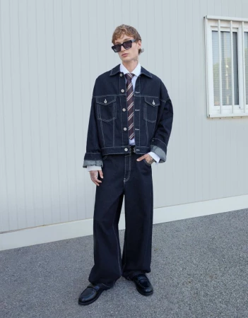 Male fashion model poses in denim jacket and wide-leg jeans with black loafers against a plain wall on an overcast day outdoors.