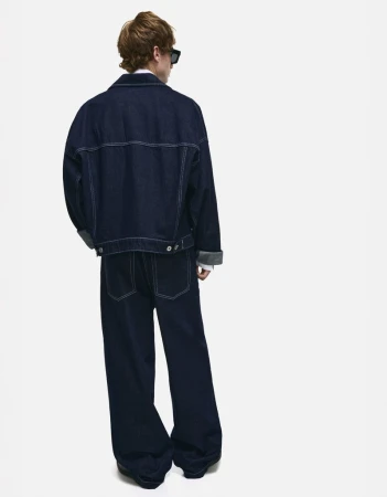 Back view of a person in a dark denim jacket and pants, turned away from the camera on a bright white seamless background.