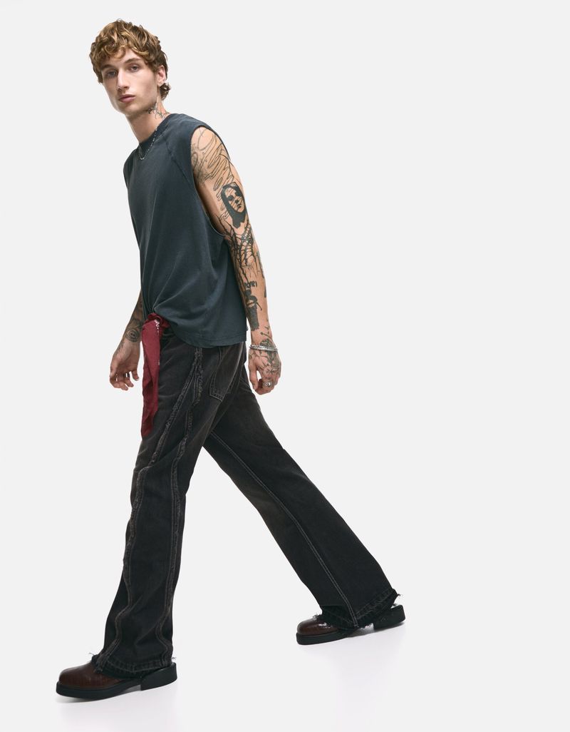 A light-skinned male model with tattoos poses in a sleeveless top and wide-leg jeans against a stark white background, showcasing fashion.