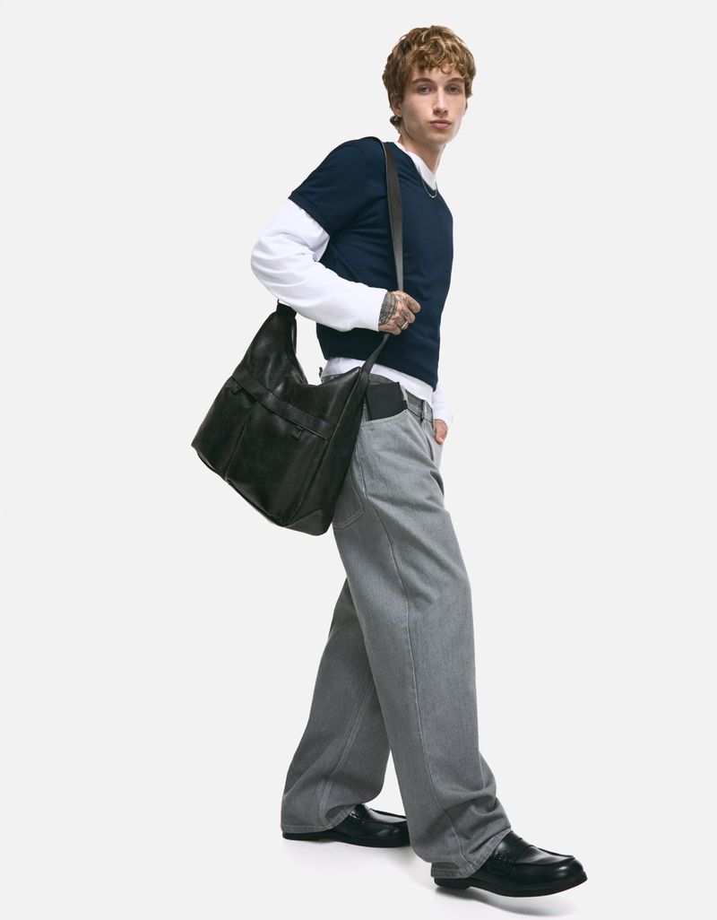 A young adult man is walking and wearing a dark sweater, gray pants, and black shoes, and carrying a large black hobo bag over his shoulder.
