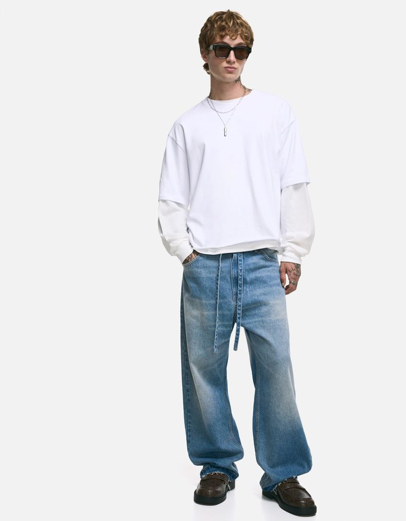 Full body shot of a young adult male with curly brown hair wearing a white layered shirt, baggy jeans, sunglasses and shoes on white.