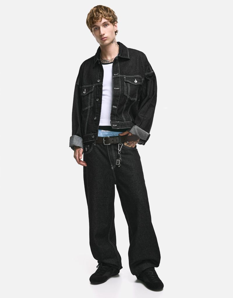 Full-length shot of a young adult man in a dark denim jacket and baggy jeans, posing against a white backdrop in a fashion-forward style.